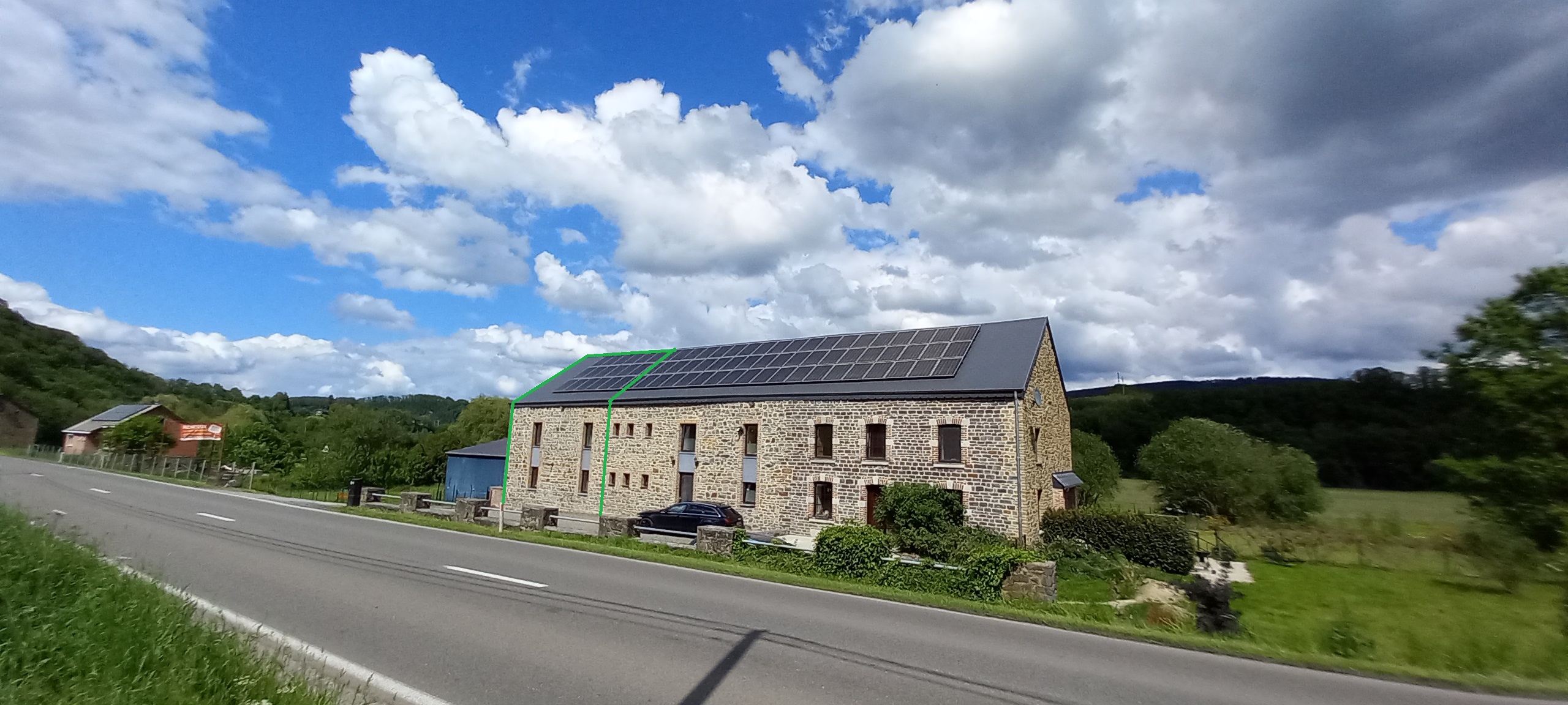 De foto van het gebouw van Amor Natura, dit betreft een gebouw met aan de buitenkant bakstenen, een zwart dak met hierop verschillende zonnepanelen. De lucht is blauw en bevat enkele wolken.